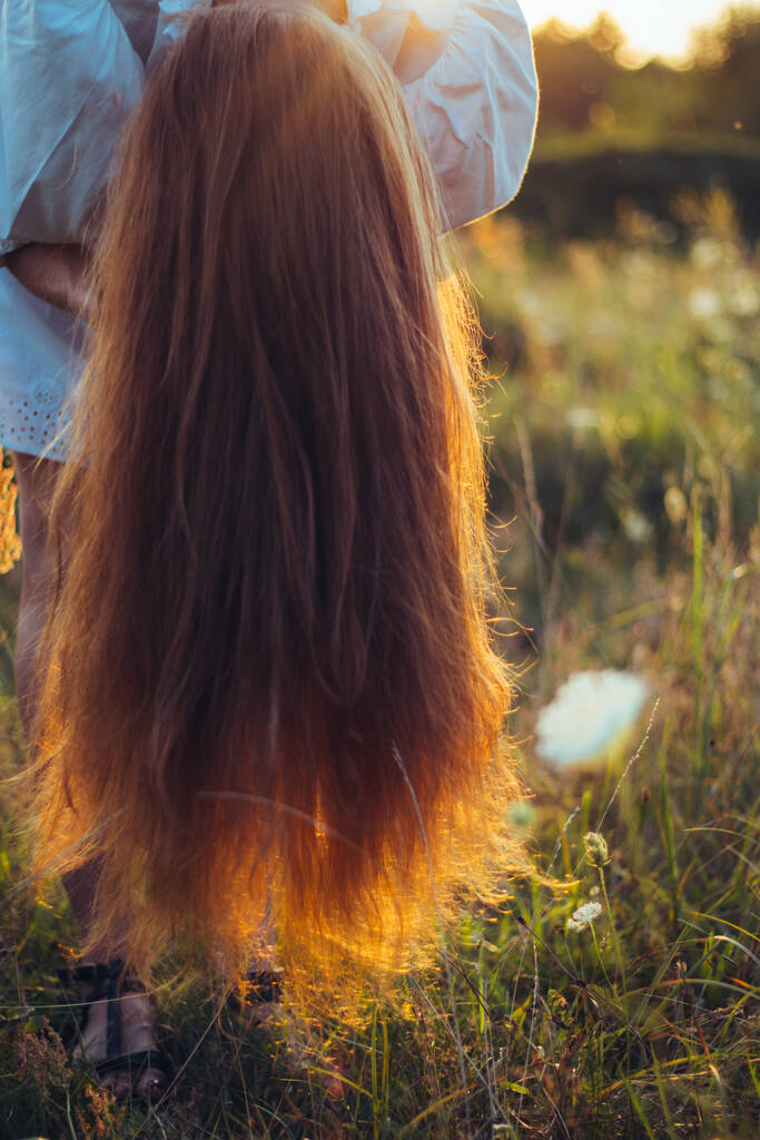 Güzel, uzun saçlı bir kadın saçını güneşli bir yaz tarlasında bağlamak için eğildi. Lüks, uzun, kırmızı, sağlıklı saçlar, doğal bakım ve bakım kavramı. Doğal gün batımı ışığı, seçici odak - Fotoğraf, Görsel