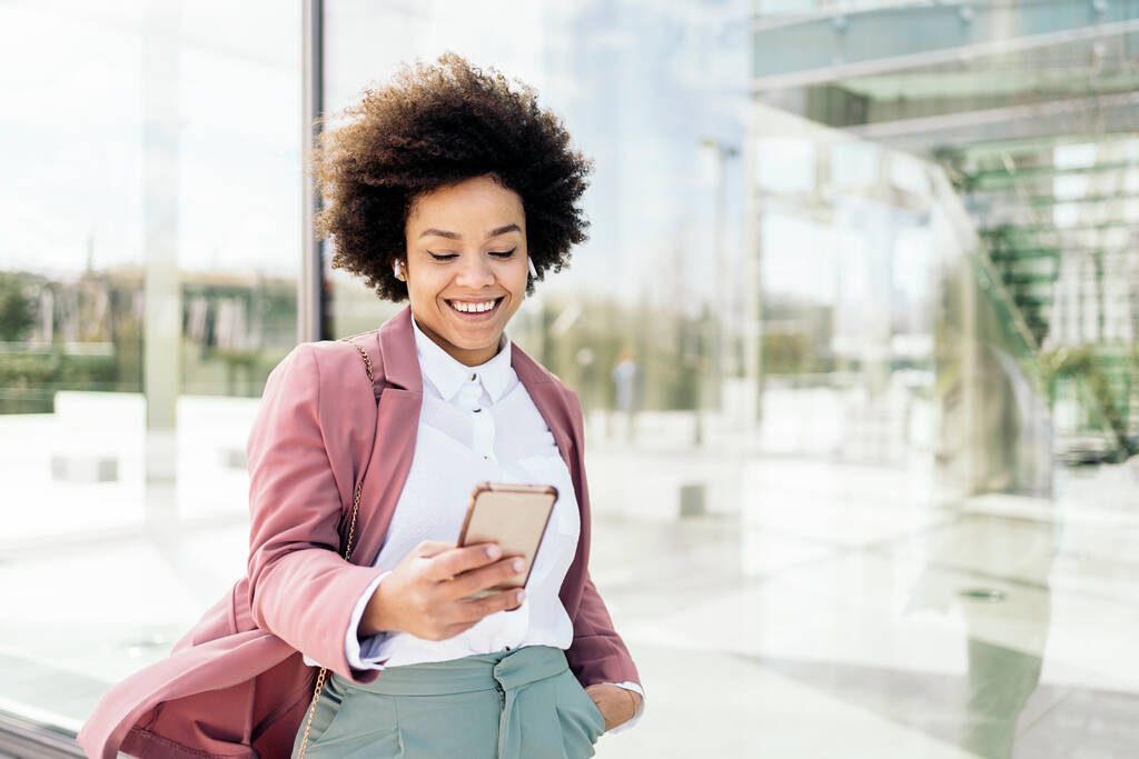 Mulher de negócios preto feliz usando seu telefone celular ao ar livre. Ela está vestindo roupas formais e usando fones de ouvido. - Foto, Imagem