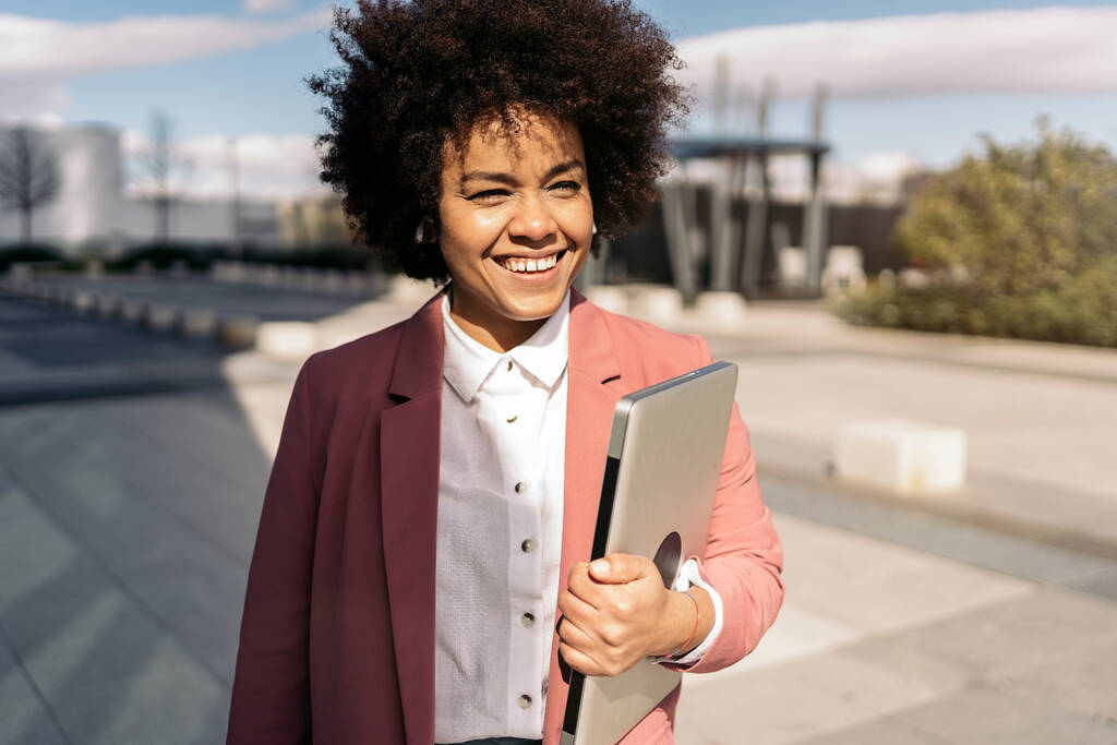 Happy black business woman with afro hair smiling and holding her laptop in the city. - Photo, Image