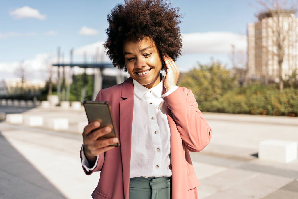 Mujer de negocios bastante negro con pelo afro utilizando su teléfono móvil al aire libre. Lleva ropa formal.. - Foto, Imagen