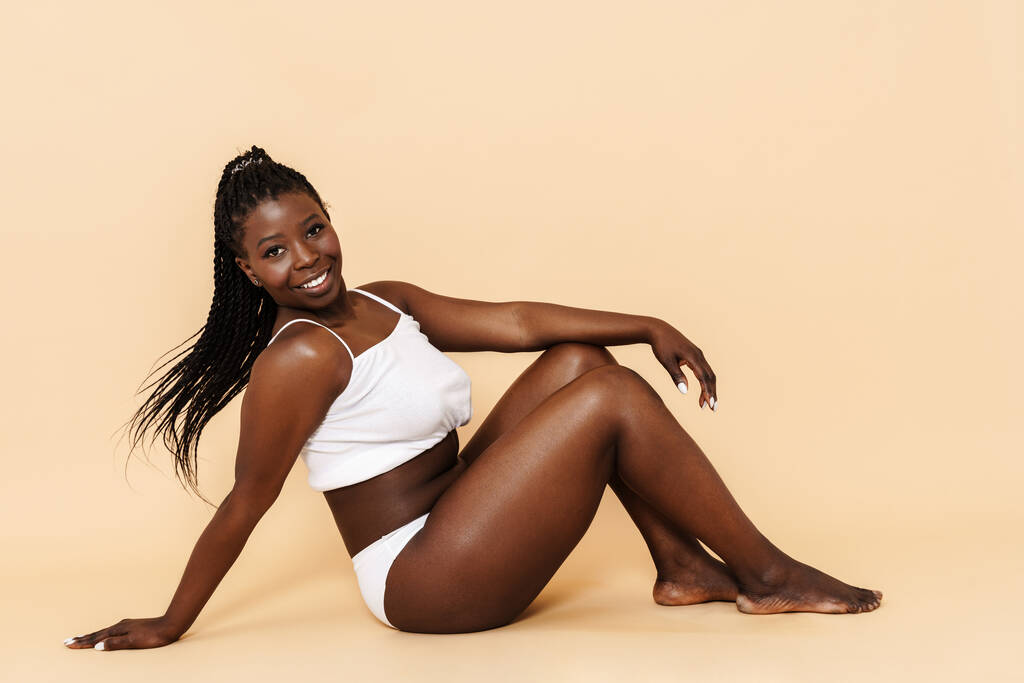 Schwarze junge Frau in weißer Unterwäsche sitzt isoliert auf dem Boden vor beigem Hintergrund - Foto, Bild