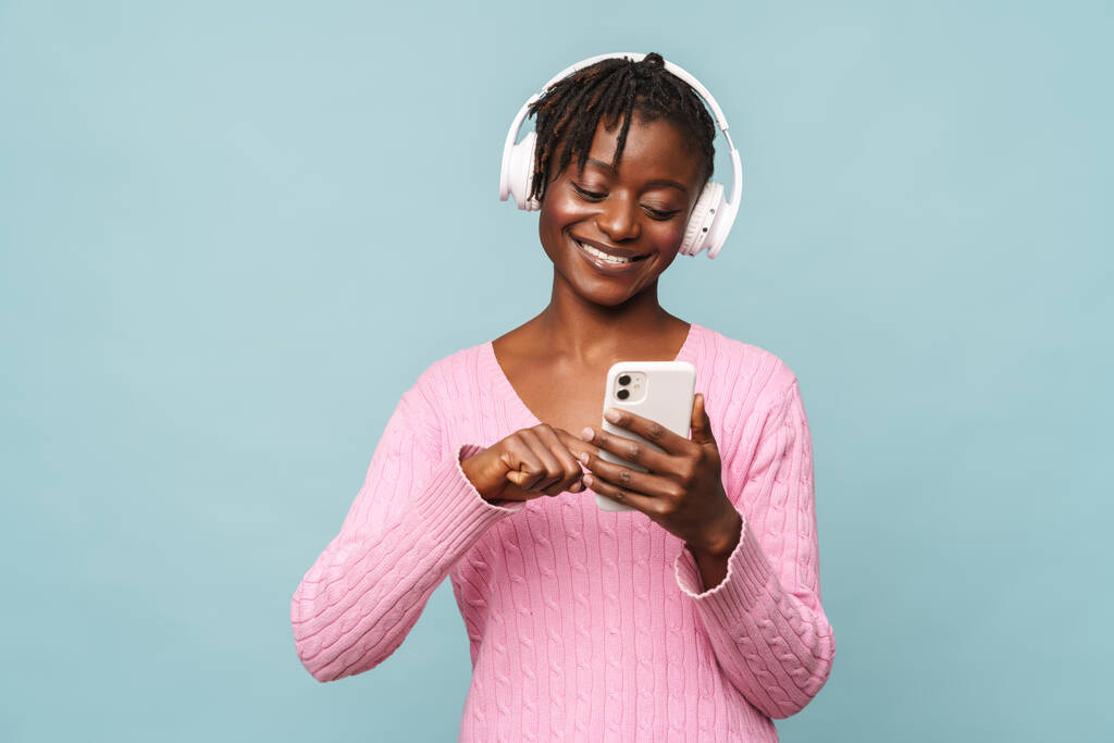 Afrykańska młoda kobieta uśmiecha się podczas korzystania z telefonu komórkowego i słuchawek odizolowanych na niebieskim tle - Zdjęcie, obraz
