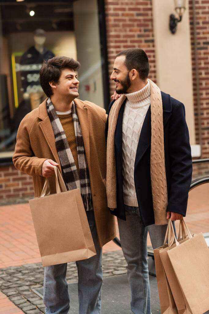 スタイリッシュな服装をしたゲイのカップルが買い物袋を持って街中で笑顔で - 写真・画像