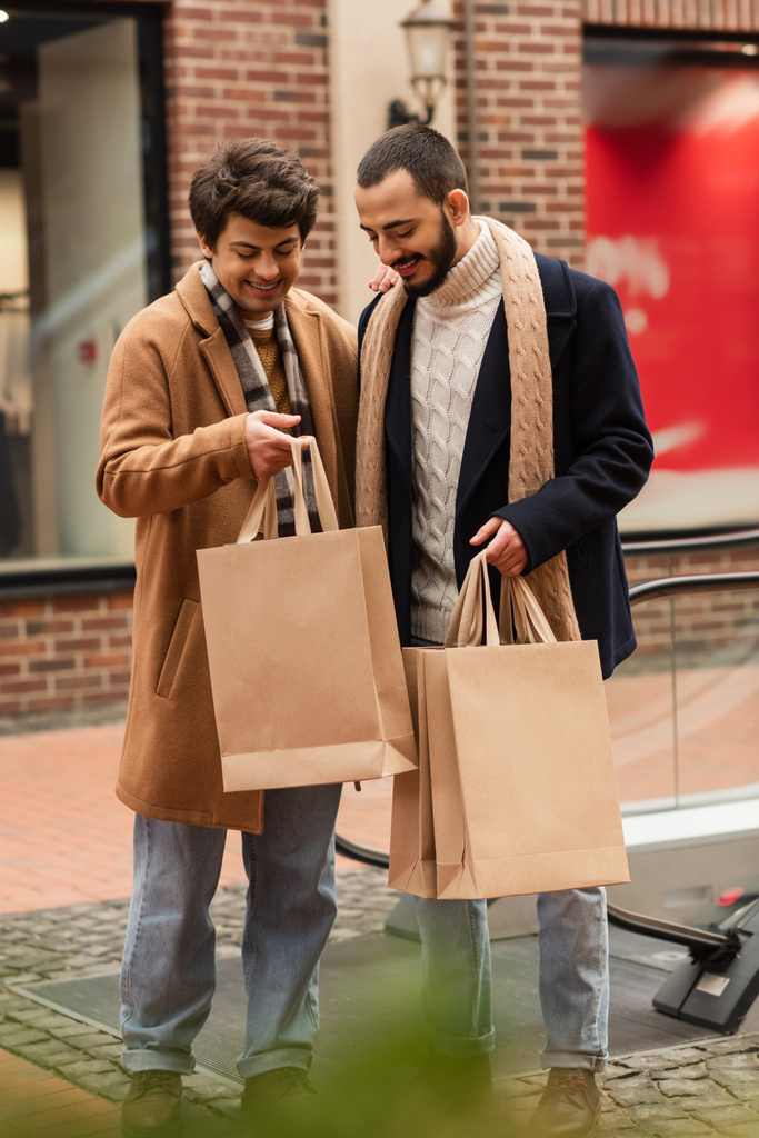 vrolijk gay paar in trendy kleding staan met winkelen tassen in de buurt wazig gebouw - Foto, afbeelding