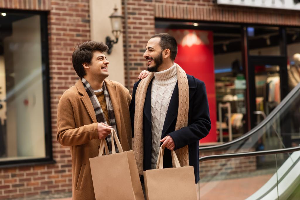 stijlvol gay paar met winkelen tassen glimlachen naar elkaar in de buurt winkel op wazig achtergrond - Foto, afbeelding