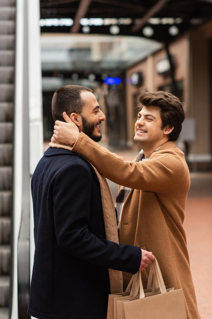 Seitenansicht eines fröhlichen schwulen Mannes, der sich an den Hals eines trendigen Freundes schmiegt, der draußen Einkaufstüten hält - Foto, Bild