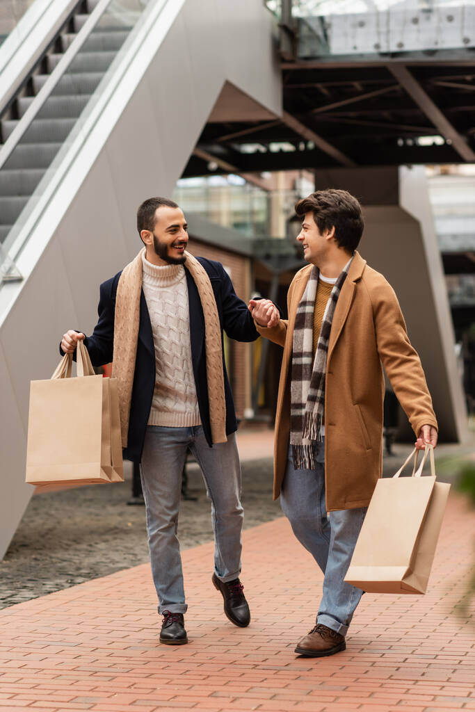 feliz gay los hombres en elegante trajes cogido de la mano mientras caminando con compras bolsas en urbano calle - Foto, imagen