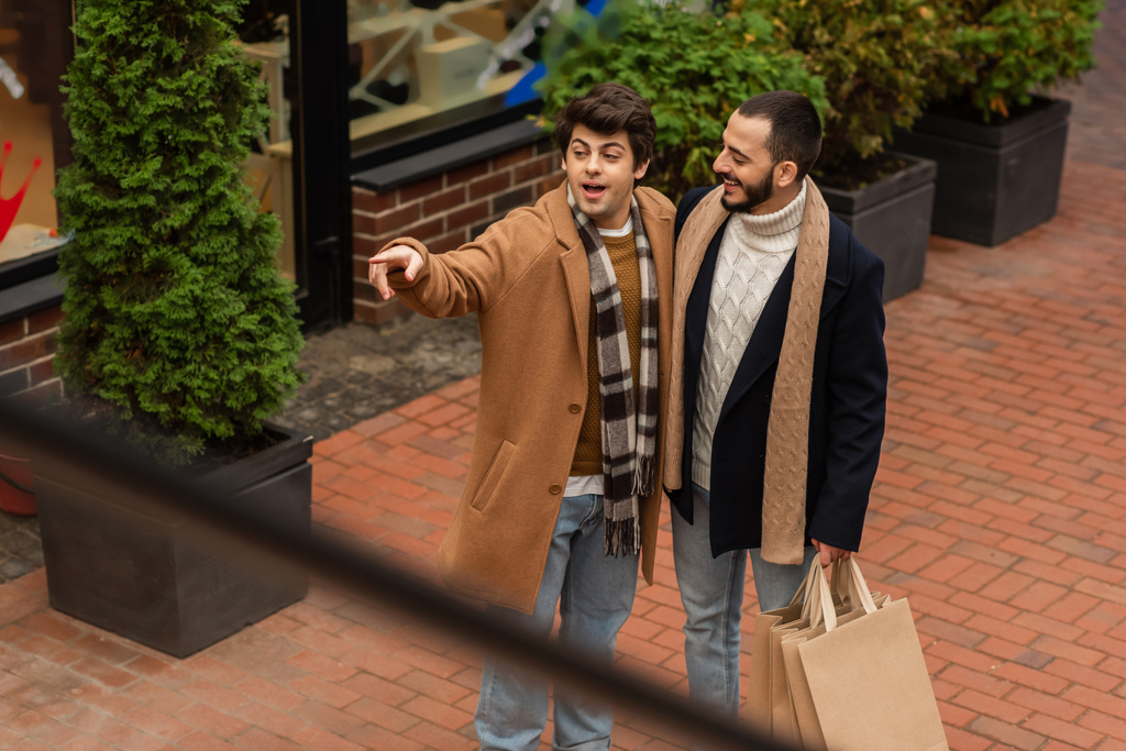 hoge hoek uitzicht van verbaasd gay man weg te kijken en wijzen met vinger in de buurt glimlachende vriend met shopping tassen  - Foto, afbeelding