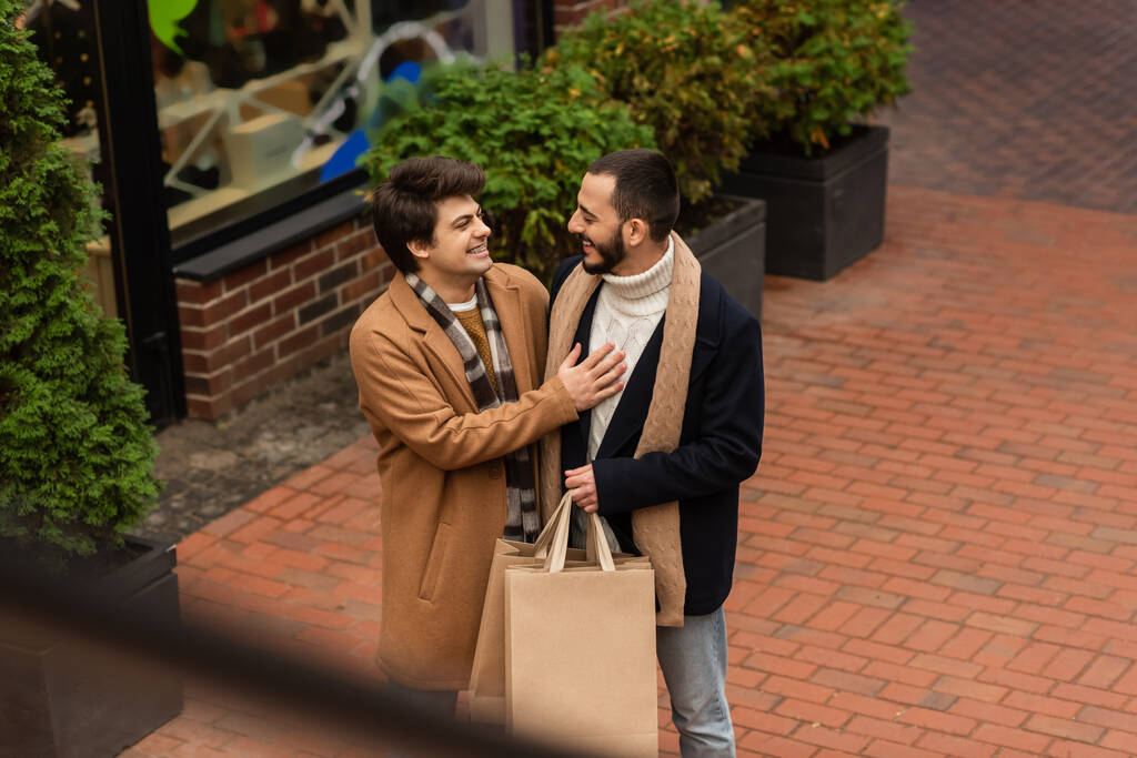 high angle view of gay man laughing near bearded boyfriend with shopping bags on urban street - Photo, Image