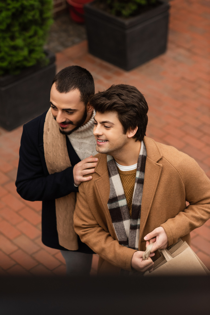 high angle view of joyful and trendy gay couple with shopping bags looking away on street - Photo, Image
