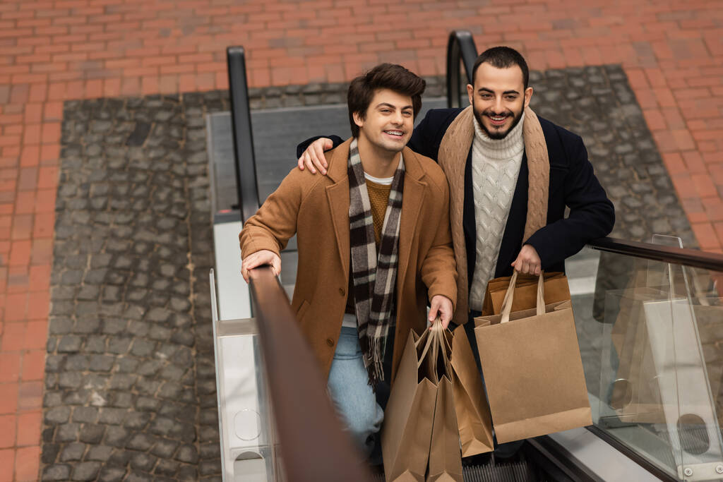 Blick aus der Vogelperspektive auf positive homosexuelle Paare in trendigen Outfits, die Einkaufstüten auf Rolltreppe halten - Foto, Bild