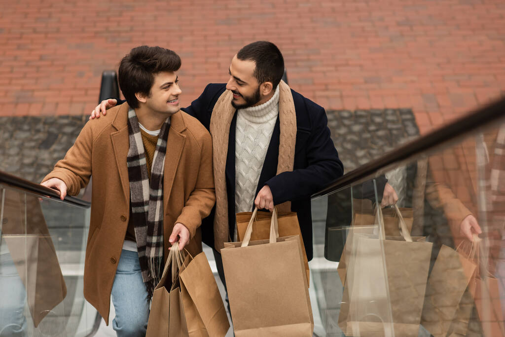 hoge hoek uitzicht van gay paar in sjaals holding shopping tassen en glimlachen naar elkaar op roltrap - Foto, afbeelding