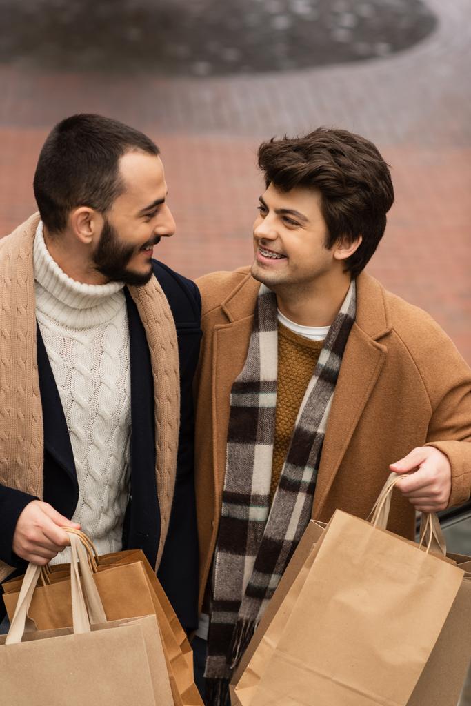 young and fashionable gay partners with shopping bags smiling at each other outdoors - Photo, Image