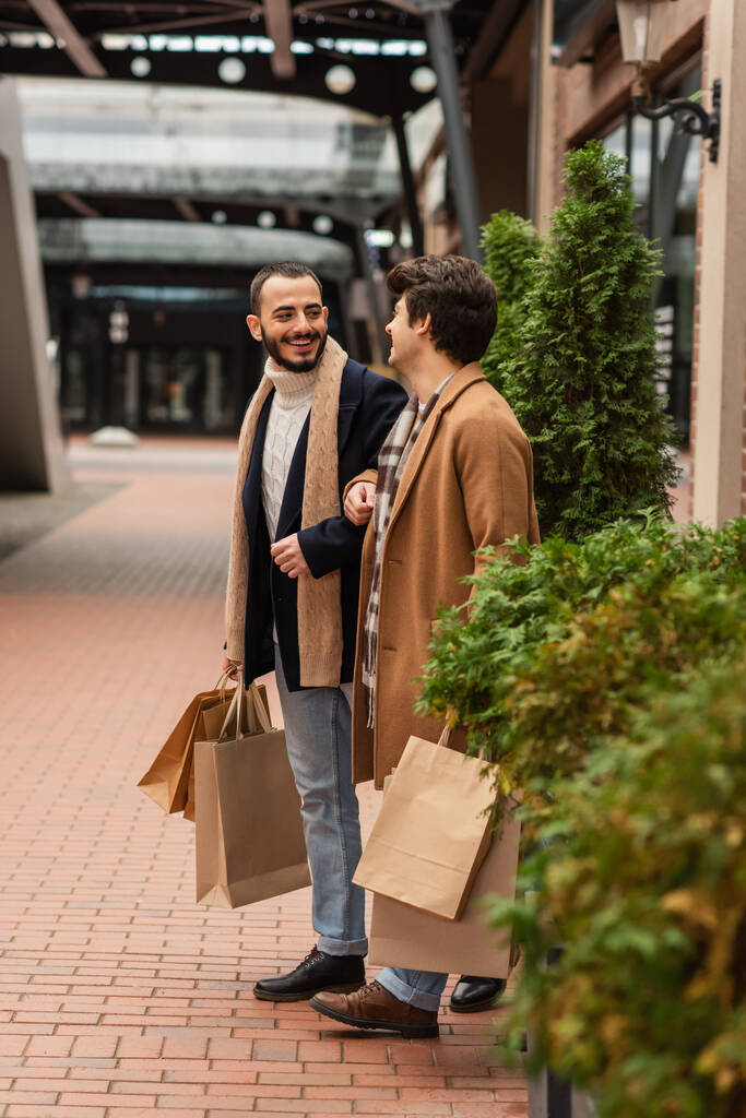 fashionable gay partners with shopping bags smiling at each other near green plants on street - Photo, Image