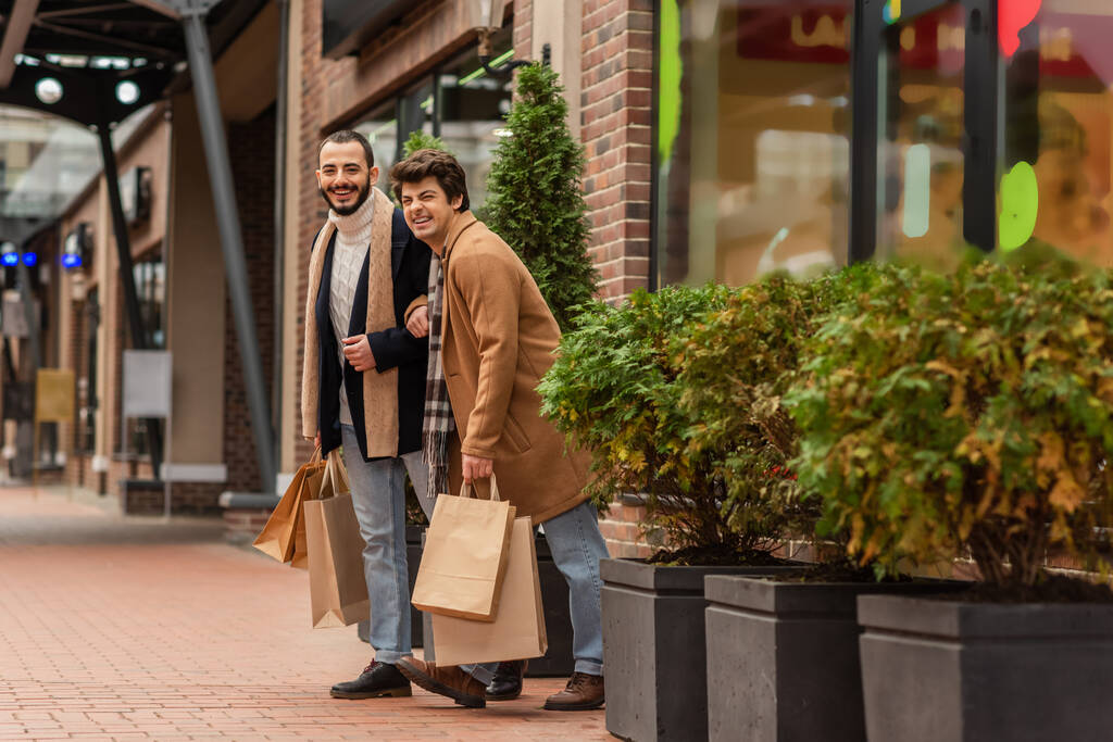 pełna długość modny i wesoły gej mężczyźni stojąc z zakupy torby w pobliżu sklep i rośliny na ulicy - Zdjęcie, obraz