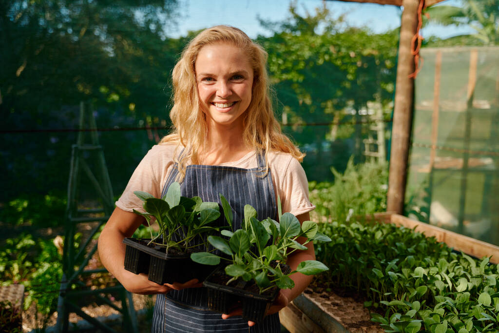 Happy young caucasian woman in apron looking at camera and holding plants in trays at plant nursery - Photo, Image