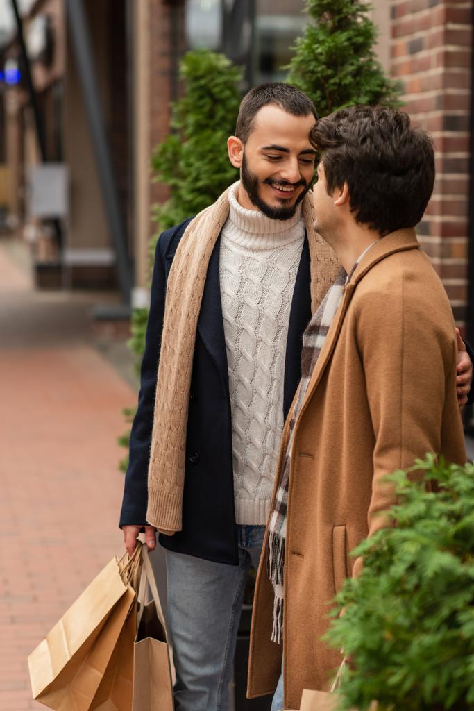 流行の衣装に身を包んだゲイの男が買い物袋を持って路上でボーイフレンドの近くで笑顔 - 写真・画像