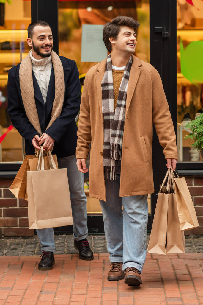 volledige lengte van trendy en gelukkig homoseksuele mannen met boodschappentassen in de buurt wazig showcase op stedelijke straat - Foto, afbeelding