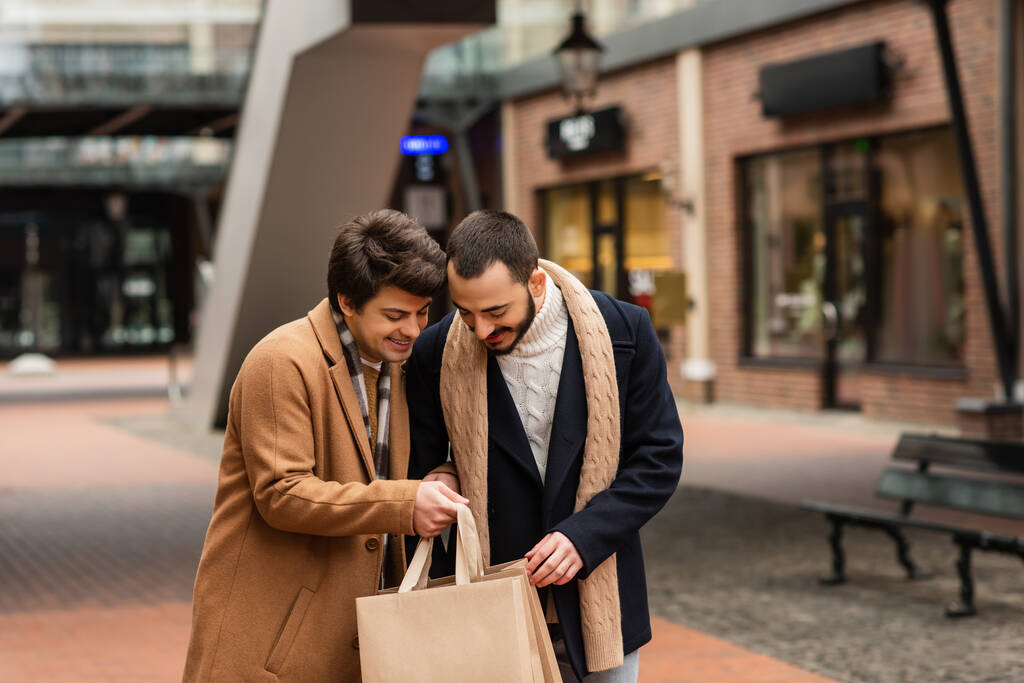 felice gay coppia in trendy vestito guardando in shopping borse su blurred strada - Foto, immagini