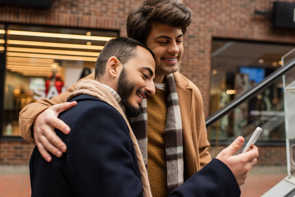 giovane e felice gay uomo in moda vestito abbracciare fidanzato con smartphone su urbano strada - Foto, immagini
