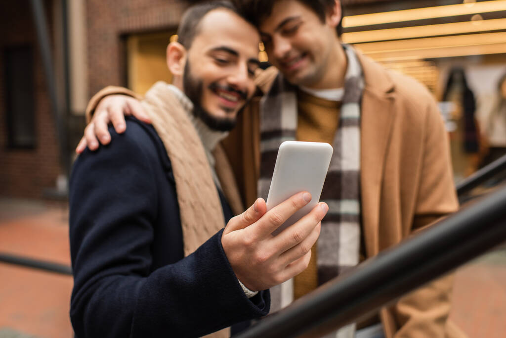 Lächelnder schwuler Mann umarmt bärtigen Freund mit Handy im Freien auf verschwommenem Hintergrund - Foto, Bild