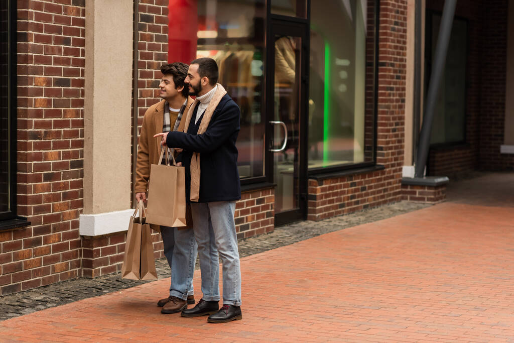 на всю длину бородатый гей с сумками для покупок, указывающими на витрину рядом с молодым парнем - Фото, изображение