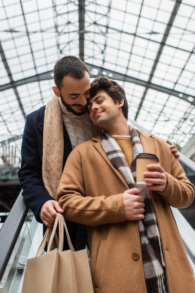 heureux gay l'homme avec café pour aller debout avec fermé les yeux près de barbu copain avec shopping sacs sur escalator - Photo, image