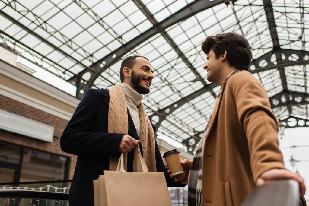 low angle view of stylish homosexual couple with shopping bags and paper cup under transparent roof outdoors - Photo, Image