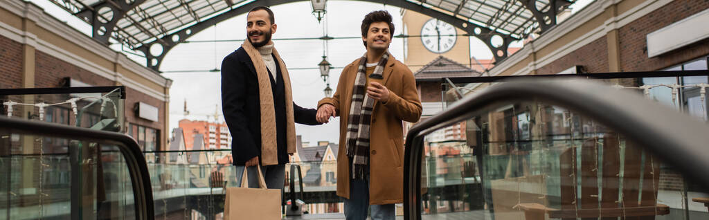 Alışveriş torbaları ve kahve taşıyan neşeli ve şık gay erkekler şehir sokaklarında el ele tutuşuyorlar. - Fotoğraf, Görsel