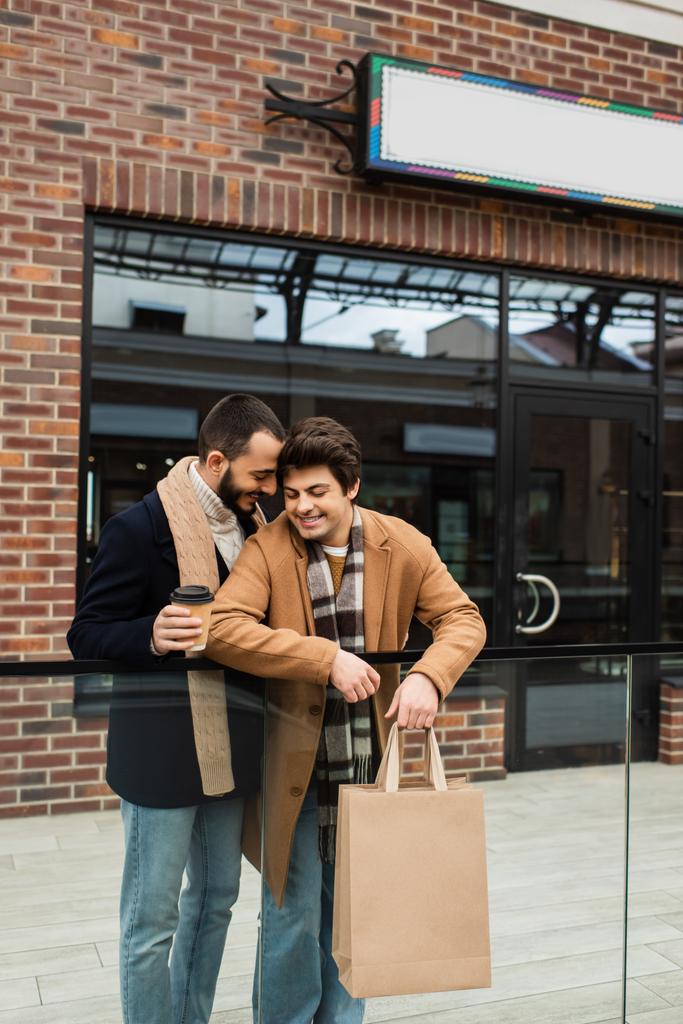 socios gay de moda con bolsas de compras y café para ir sonriendo cerca de valla de vidrio y edificio con escaparate - Foto, imagen