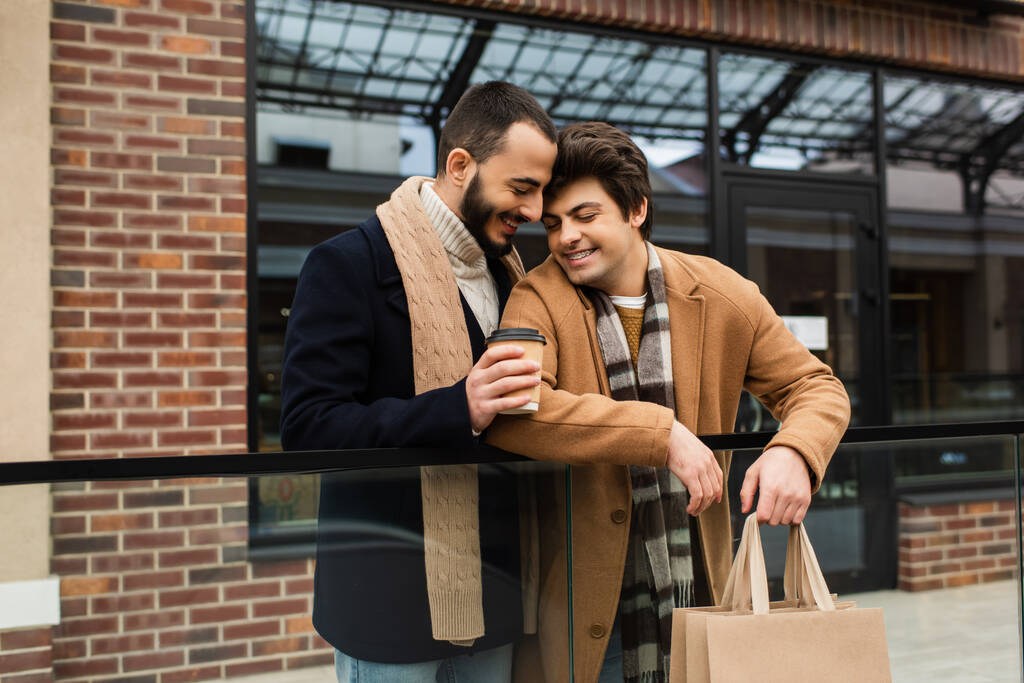 gelukkig gay man holding takeaway drankje in de buurt jong vriendje met winkelen tassen en winkel op de achtergrond - Foto, afbeelding