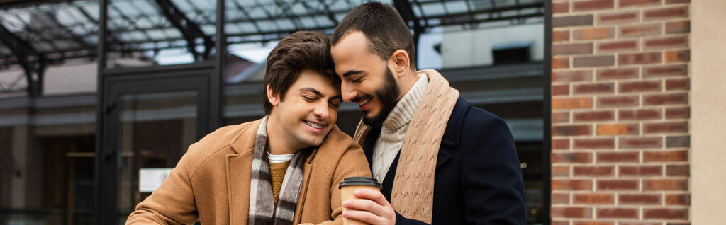fröhlicher bärtiger Mann hält Kaffee in der Nähe des trendigen homosexuellen Partners auf der Straße der Stadt, Banner - Foto, Bild