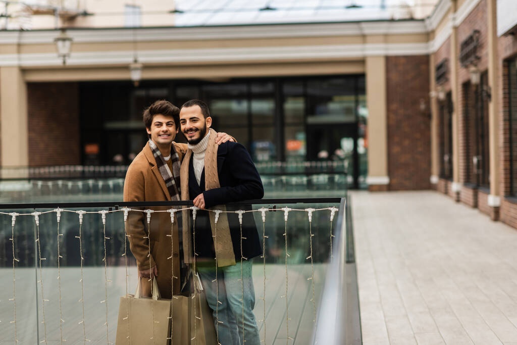 μοντέρνα και ευτυχισμένη ομοφυλόφιλους άνδρες με τσάντες ψώνια στέκεται κοντά σε γυάλινο φράχτη και θολή κτίριο - Φωτογραφία, εικόνα