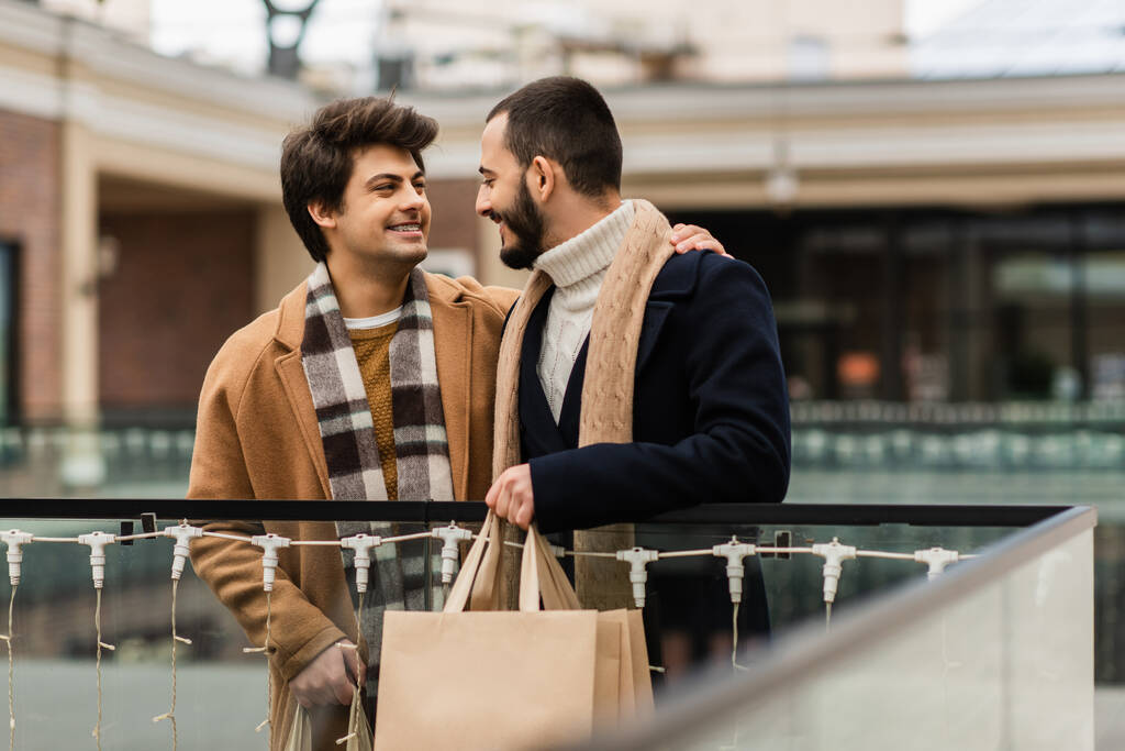 szczęśliwy geje w modne płaszcze i szaliki trzymając torby na zakupy i patrząc na siebie na miejskiej ulicy - Zdjęcie, obraz