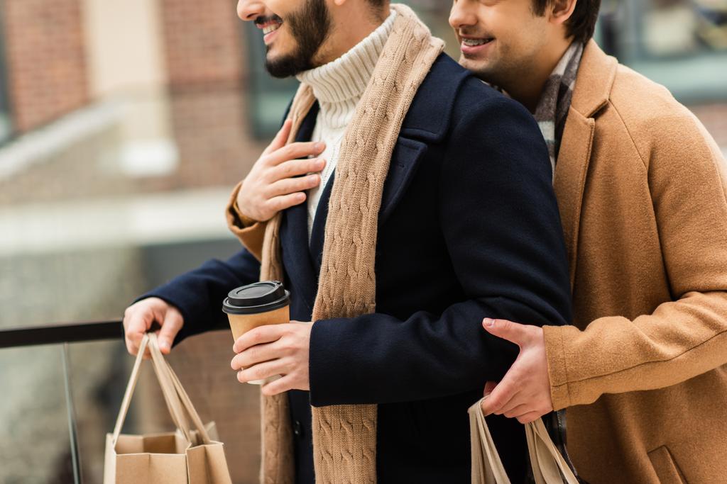 καλλιεργημένη άποψη του χαμογελαστού gay άντρας αγκαλιάζει γενειοφόρο φίλο με καφέ για να πάει και τσάντες ψώνια - Φωτογραφία, εικόνα