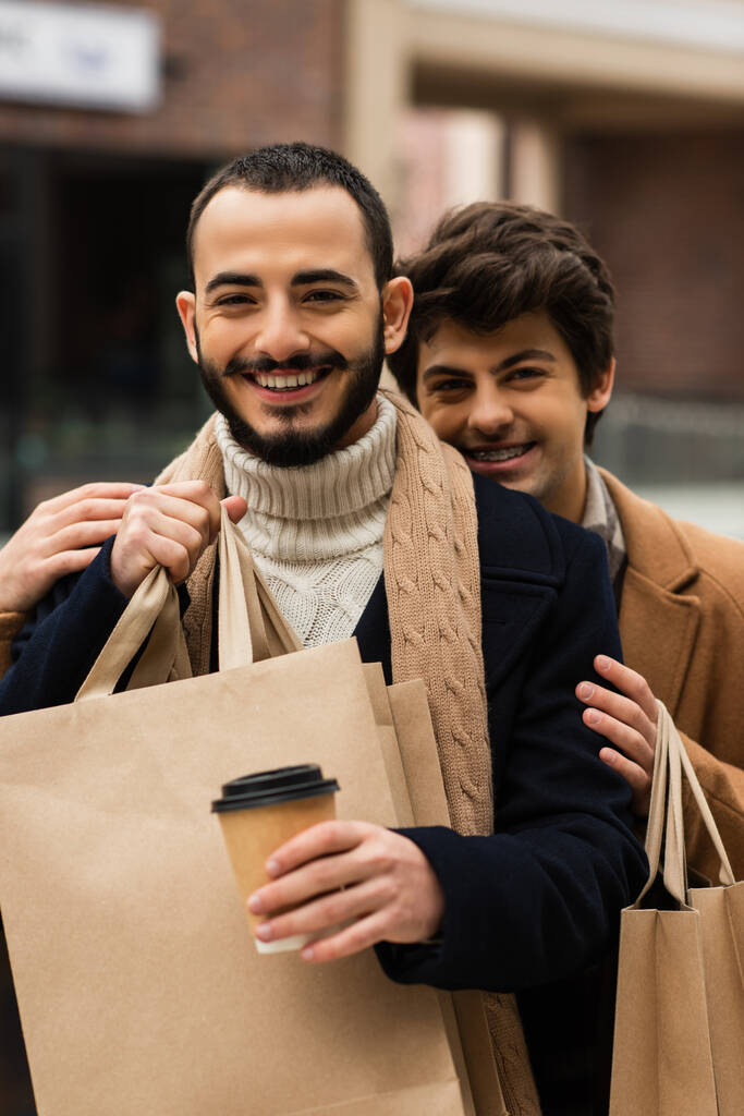 veselý gay muž objímající vousatý přítel držící papírový pohár a nákupní tašky - Fotografie, Obrázek