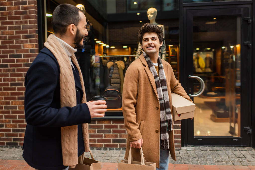 Lächelnder schwuler Mann mit Einkäufen, der seinen Freund mit Pappbecher in der Nähe des Geschäfts an der Stadtstraße anschaut - Foto, Bild