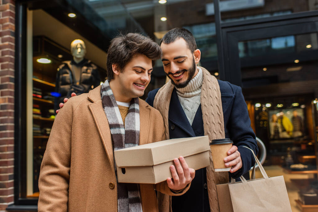 hymyilevä parrakas mies kahvia mennä katsomaan kenkälaatikko kädessä poikaystävä lähellä hämärtynyt näyteikkuna kadulla - Valokuva, kuva