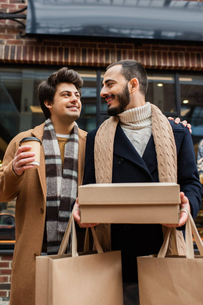 heureux gay couple dans branché tenues regarder l 'autre tandis que debout avec achats et à emporter boisson - Photo, image