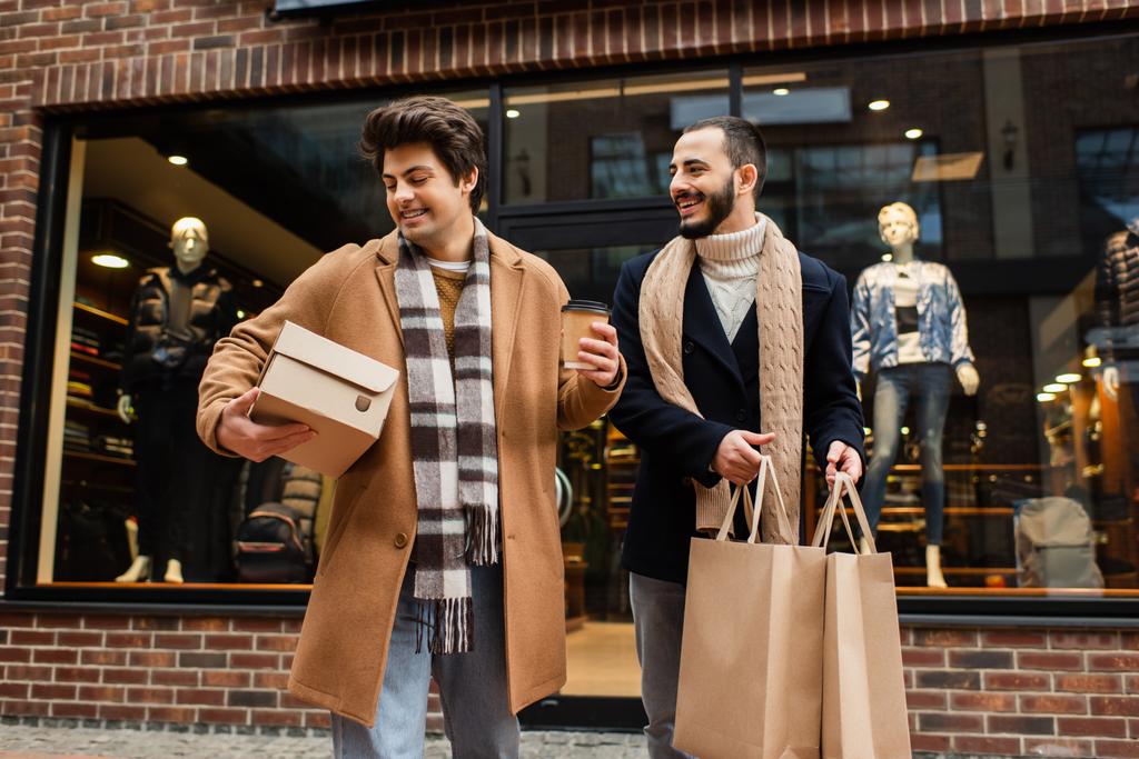 trendi meleg férfi bevásárló táskák nézi vidám barátja cipős doboz és papír pohár közelében kirakat  - Fotó, kép