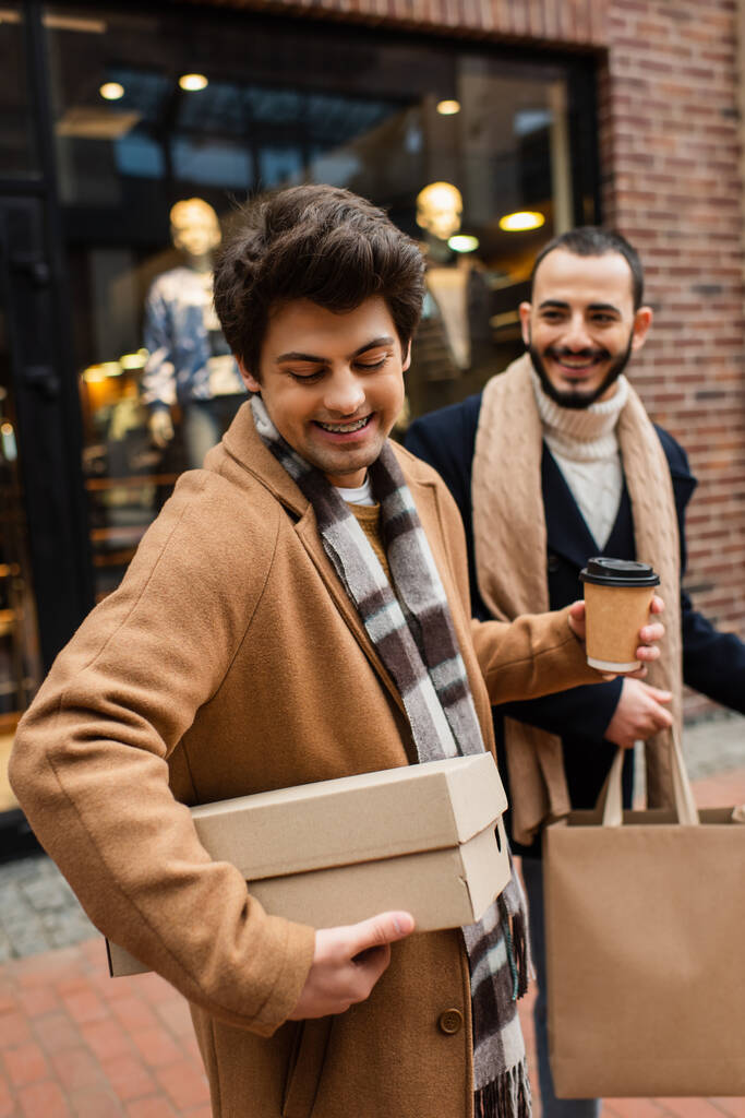 веселий гей чоловік дивиться на взуття поблизу усміхненого хлопця з сумкою для покупок і паперовою чашкою на розмитій вітрині
  - Фото, зображення