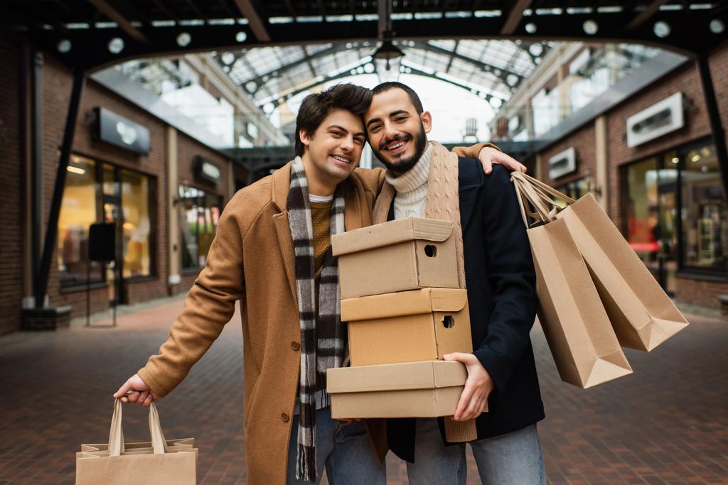 pleased gay man with shopping bags hugging bearded boyfriend with shoeboxes on urban street - Photo, Image