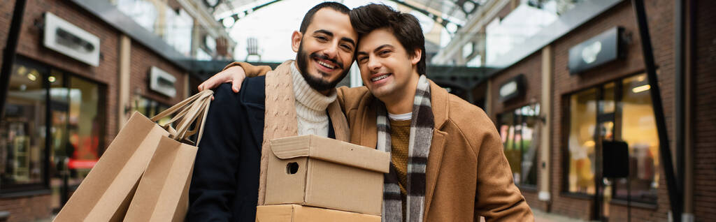 alegre hombre con bolsas de compras abrazando pareja gay con cajas de zapatos cerca de las tiendas borrosas en el fondo, pancarta - Foto, Imagen
