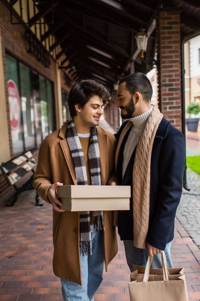 young gay man in checkered scarf holding shoebox near smiling boyfriend with shopping bags - Photo, Image