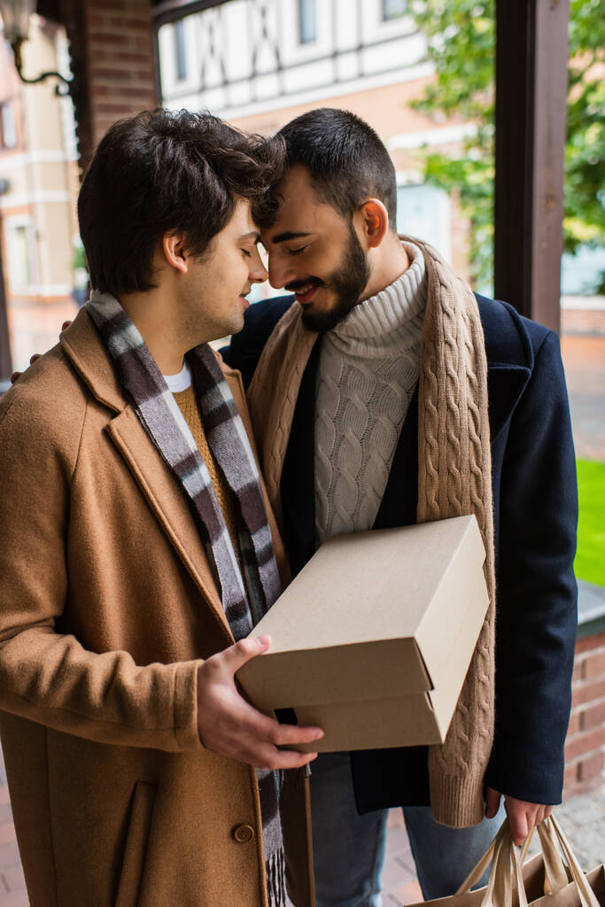 mladý gay muž se zavřenýma očima drží krabice od bot, zatímco stojí tváří v tvář se šťastným vousatým přítelem  - Fotografie, Obrázek