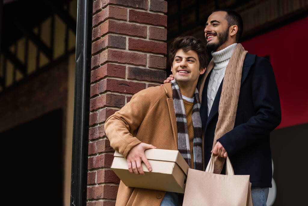 alegre y elegante pareja gay con bolsas de compras y caja de zapatos mirando lejos cerca de la columna de ladrillo - Foto, Imagen