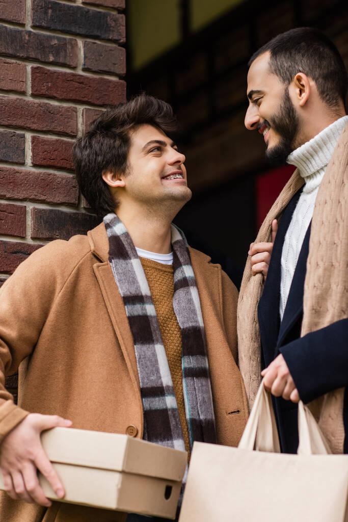alegre y de moda pareja gay con compras sonriendo el uno al otro cerca de ladrillo pared al aire libre - Foto, Imagen