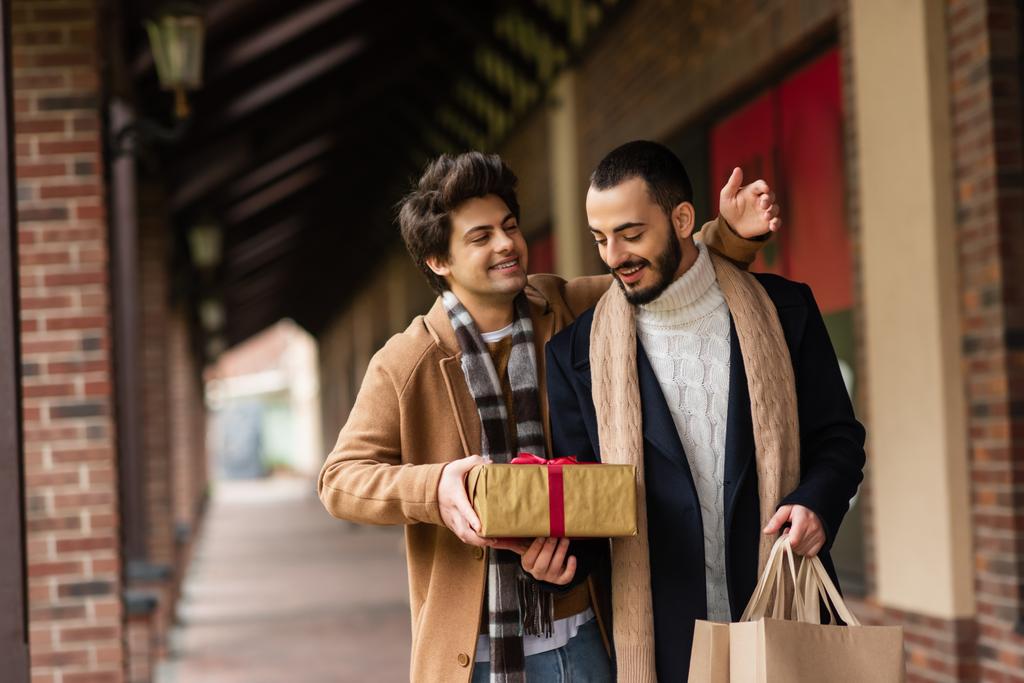 alegre gay hombre presentando regalo caja a complacido y de moda novio celebración bolsas de compras en la ciudad calle - Foto, imagen