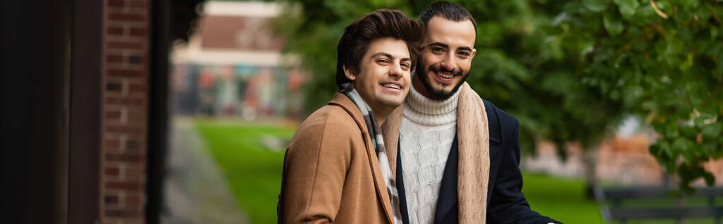 молодая и модная гей-пара в пальто в шарфах, улыбающаяся в камеру на улице, баннер - Фото, изображение