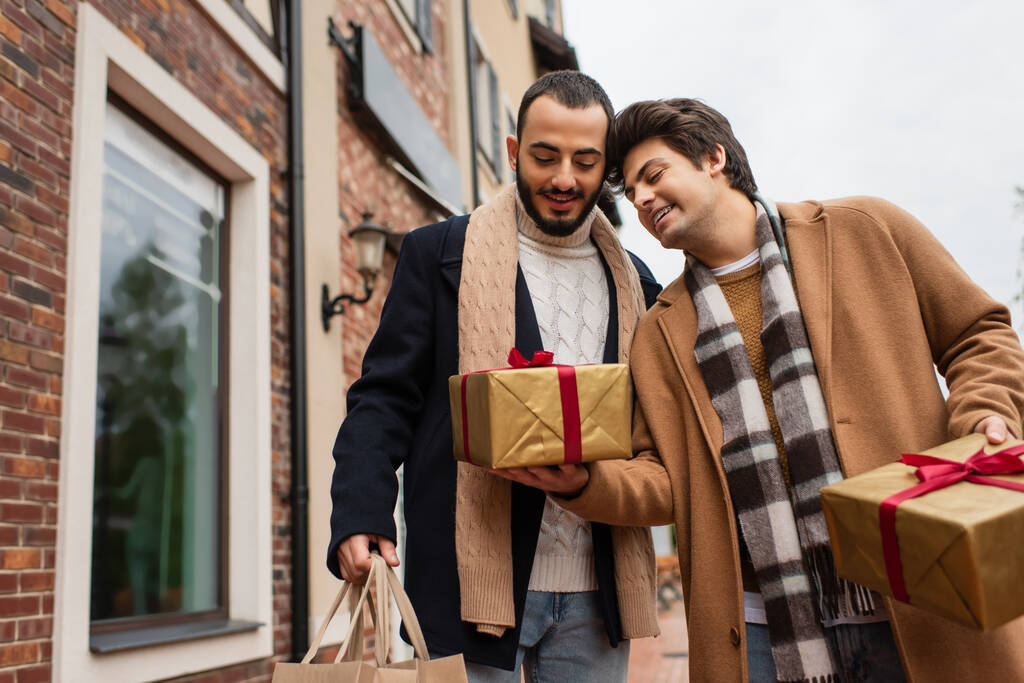 νεαρός ομοφυλόφιλος άνδρας κρατώντας κουτιά δώρων κοντά χαρούμενος και μοντέρνος φίλος με τσάντες ψώνια στην αστική δρόμο - Φωτογραφία, εικόνα
